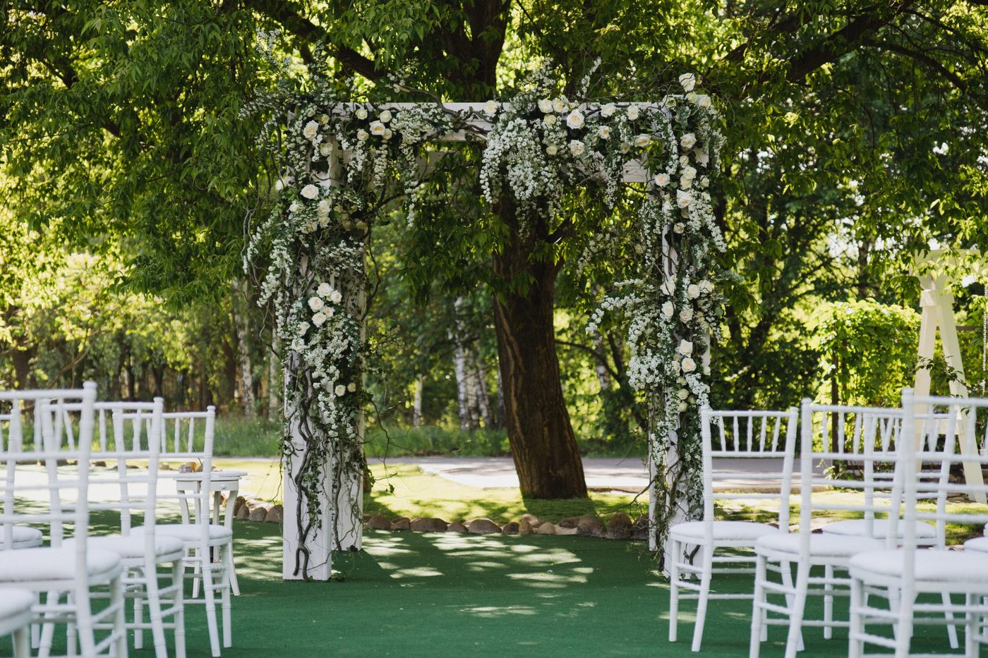 A white flower-covered wedding arch under a tree, with white chairs on either side